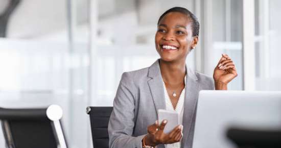 African black business woman using smartphone while working on laptop at office. Smiling mature african american businesswoman lookingup at copy space while working on phone. Successful woman entrepreneur think about new business ideas.
